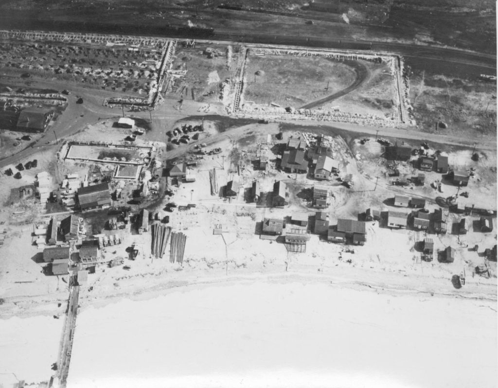 Aerial photograph of shacks on a frozen shoreline, train tracks, and construction.