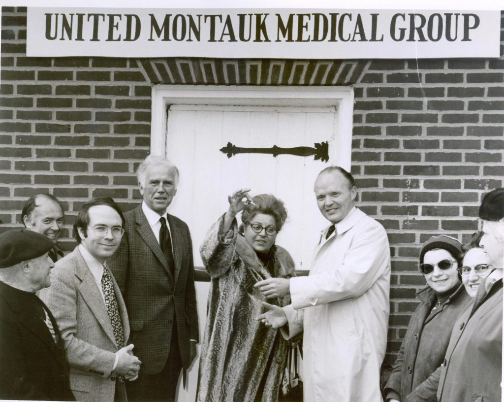 Five people stand in front of a brick building with a sign saying United Montauk Medical Center. In the center a woman in a fur coats holds up a set of keys.