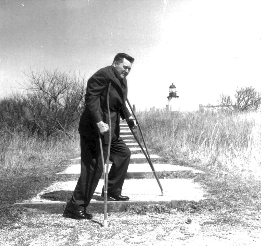 A man with crutches stands at the bottom of a series of steps with the Montauk Lighthouse behind him.