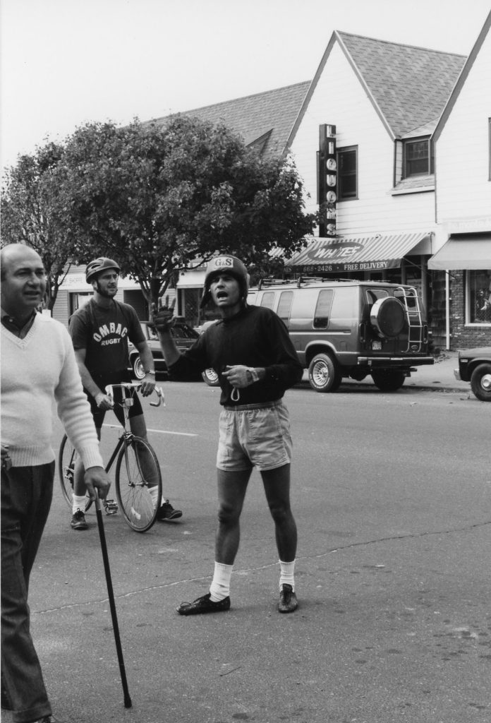 Man in shorts and helmet stands on road instructing bicyclists at start of race.