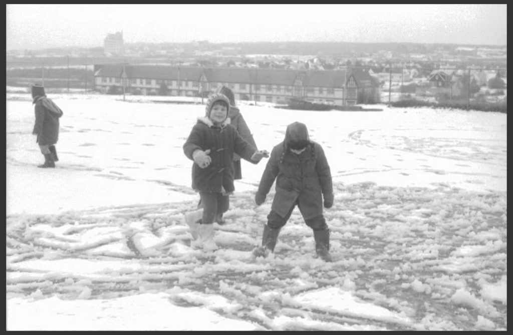 Four school aged children photographed playing in two inches of snow in an open field in front of a neighborhood.