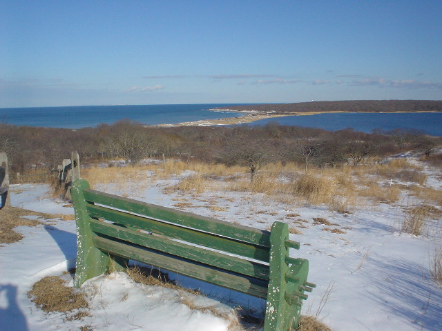 A bench overlooks flat grassy land and blue water. Snow is on the ground.