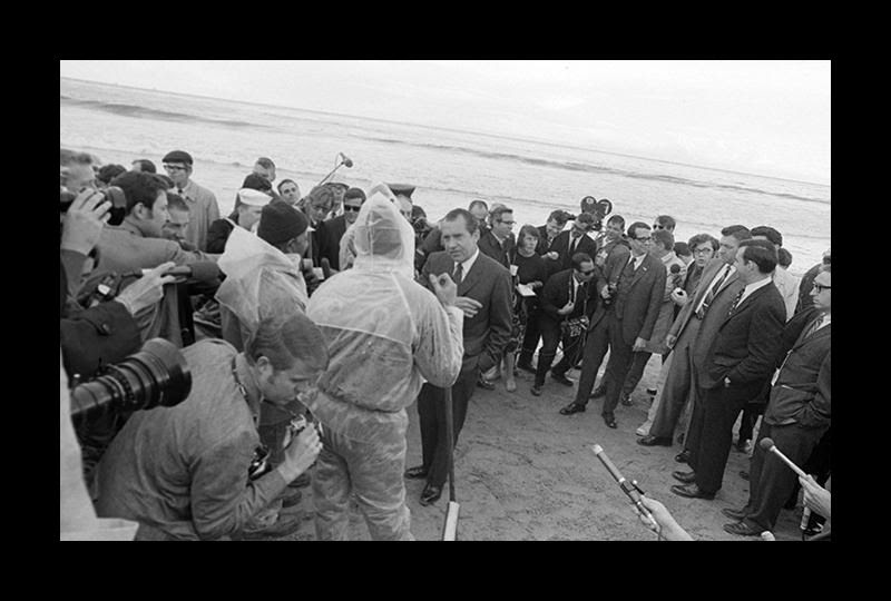 A crowd of men in suits on the ocean shore with President Nixon at center, cameramen, and two men in Haz-Mat suits.