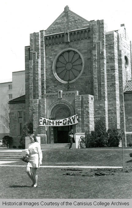 A church-like college biulding with an Earth Day banner across the front entrance and a young woman in 1960s-1970s-style miniskirt carrying books walking away in front.