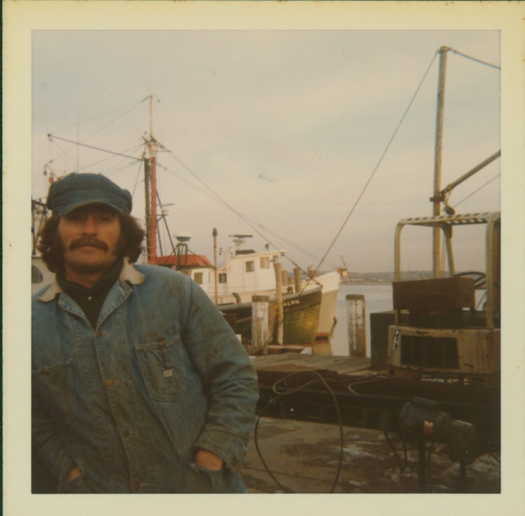 Man in denim jacket and hat with mustache posing in front of boats docked in a harbor. 