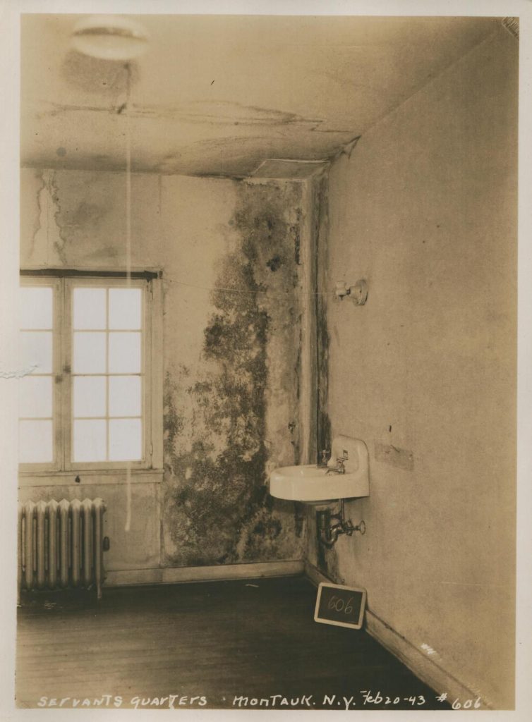 A black and white photo of the corner of a room with a window, radiator, and sink, extensive water damage and mold in the corner and on ceiling.