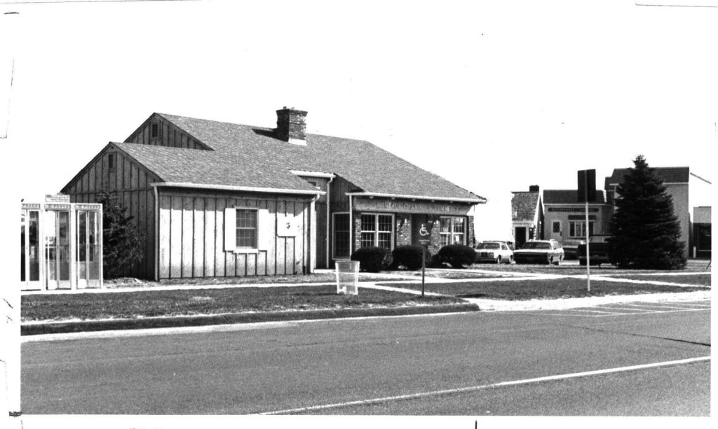 Larger Montauk Chamber of Commerce building with phone booth bank and metal trash pail outside.