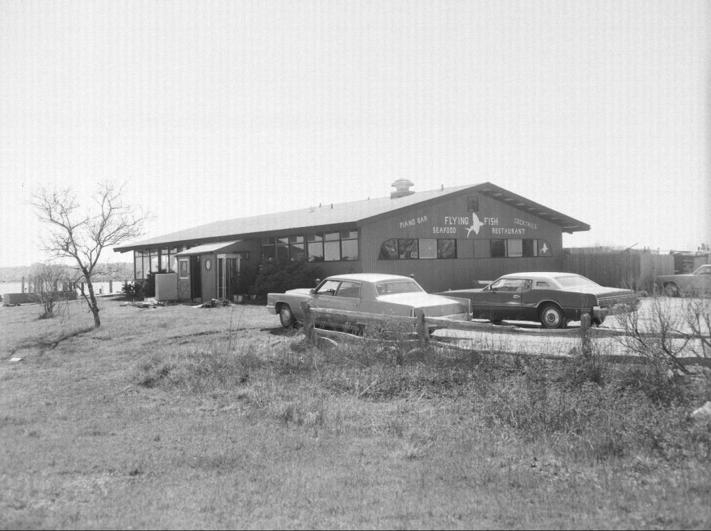 Waterfront bar and restaurant, circa 1970s, with vintage cars parked outside.