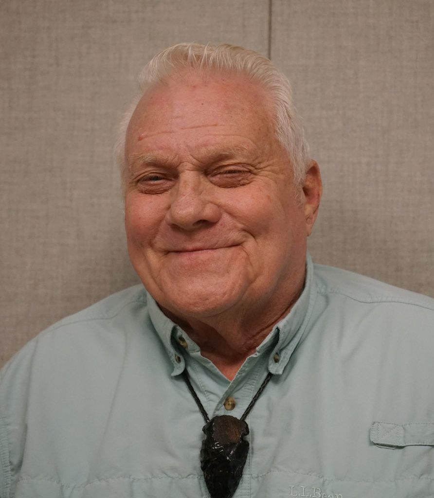 A portrait of older man from the shoulders up has white hair and smiles at the camera wearing a blue shirt. 