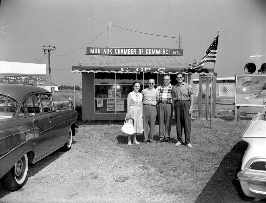 Small Montauk Chamber of Commerce building, circa 1960s, with four people posting outside and flag flying next to phone booth.