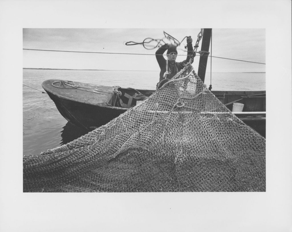 A man on a small boat on the shore hanging up a net to dry.